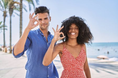 Téléchargez les photos : Jeune couple interracial à l'extérieur par une journée ensoleillée souriant positif faire ok signe avec la main et les doigts. expression réussie. - en image libre de droit