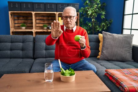 Foto de Hombre mayor con cabello gris comiendo ensalada y manzana verde con la mano abierta haciendo stop sign con expresión seria y segura, gesto de defensa - Imagen libre de derechos