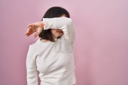 Téléchargez les photos : Femme avec le syndrome du duvet debout sur fond rose couvrant les yeux avec le bras, l'air grave et triste. concept de non-visibilité, de dissimulation et de rejet - en image libre de droit