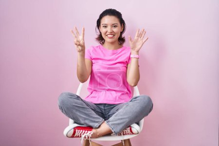 Photo for Hispanic young woman sitting on chair over pink background showing and pointing up with fingers number eight while smiling confident and happy. - Royalty Free Image