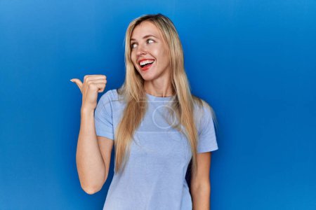 Foto de Beautiful blonde woman wearing casual t shirt over blue background smiling with happy face looking and pointing to the side with thumb up. - Imagen libre de derechos