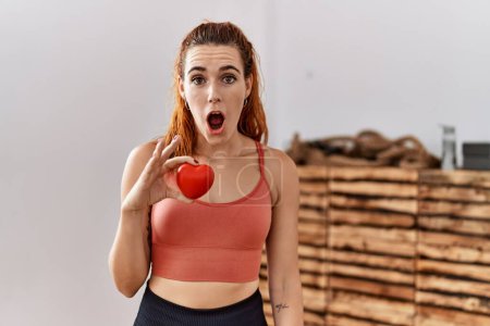 Photo for Young redhead woman holding red heart at the gym scared and amazed with open mouth for surprise, disbelief face - Royalty Free Image