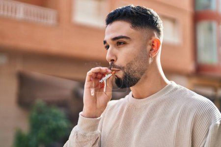 Foto de Hombre árabe joven fumando con expresión relajada en la calle - Imagen libre de derechos