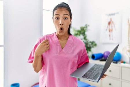 Foto de Young hispanic physiotherapist woman using computer laptop at medical clinic scared and amazed with open mouth for surprise, disbelief face - Imagen libre de derechos