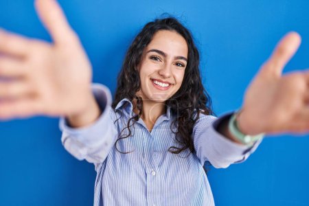 Téléchargez les photos : Jeune femme brune debout sur fond bleu regardant la caméra souriant à bras ouverts pour se faire câliner. expression joyeuse embrassant le bonheur. - en image libre de droit
