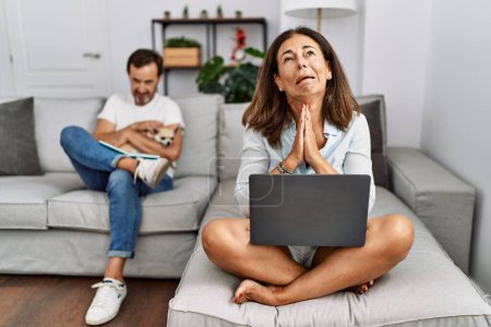 Photo for Hispanic middle age couple at home, woman using laptop begging and praying with hands together with hope expression on face very emotional and worried. begging. - Royalty Free Image