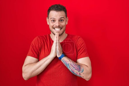 Téléchargez les photos : Jeune homme hispanique debout sur fond rouge priant avec les mains ensemble demandant pardon souriant confiant. - en image libre de droit