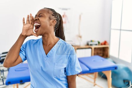 Téléchargez les photos : Femme noire avec des tresses travaillant à la clinique de récupération de la douleur criant et criant fort à côté avec la main sur la bouche. concept de communication. - en image libre de droit
