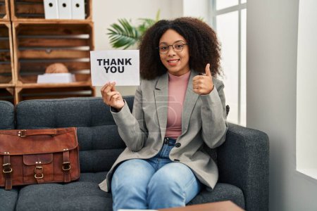 Photo for Young african american woman working at consultation office holding thank you banner smiling happy and positive, thumb up doing excellent and approval sign - Royalty Free Image