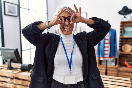 Photo for Middle age grey-haired woman working as manager at retail boutique doing ok gesture like binoculars sticking tongue out, eyes looking through fingers. crazy expression. - Royalty Free Image