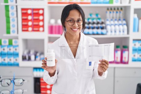 Téléchargez les photos : Jeune pharmacienne hispanique tenant une bouteille de pilules et du papier sur ordonnance à la pharmacie - en image libre de droit