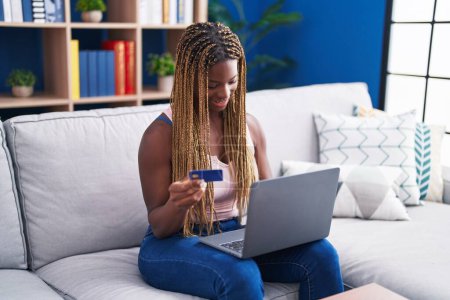 Téléchargez les photos : Femme afro-américaine utilisant ordinateur portable et carte de crédit assis sur le canapé à la maison - en image libre de droit