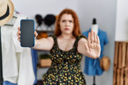Téléchargez les photos : Young redhead woman at retail shop showing smartphone screen with open hand doing stop sign with serious and confident expression, defense gesture - en image libre de droit