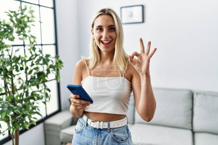 Téléchargez les photos : Jeune femme blonde en utilisant smartphone dactylographier message à la maison faire ok signe avec les doigts, sourire geste amical excellent symbole - en image libre de droit