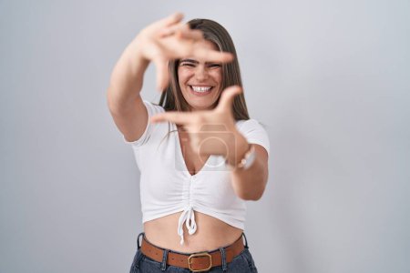 Téléchargez les photos : Jeune belle femme portant casual t-shirt blanc souriant faisant cadre avec les mains et les doigts avec le visage heureux. concept de créativité et photographie. - en image libre de droit