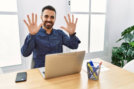 Photo for Young hispanic man with beard working at the office with laptop showing and pointing up with fingers number ten while smiling confident and happy. - Royalty Free Image