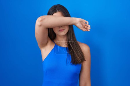 Photo for Hispanic woman standing over blue background covering eyes with arm, looking serious and sad. sightless, hiding and rejection concept - Royalty Free Image