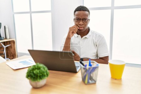 Téléchargez les photos : Jeune homme africain travaillant au bureau en utilisant ordinateur portable bouche et lèvres fermées comme zip avec les doigts. secret et silencieux, tabou parlant - en image libre de droit