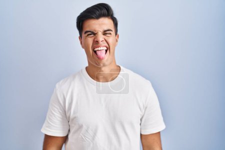 Foto de Hombre hispano de pie sobre fondo azul sacando la lengua feliz con expresión divertida. concepto de emoción. - Imagen libre de derechos