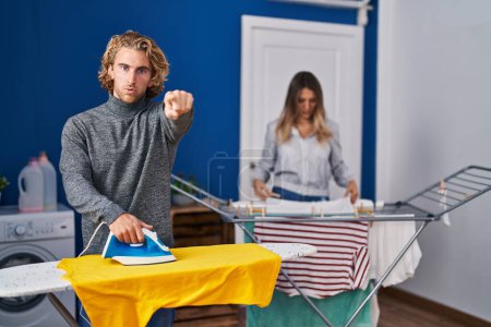 Téléchargez les photos : Couple ironing clothes at laundry room pointing with finger to the camera and to you, confident gesture looking serious - en image libre de droit