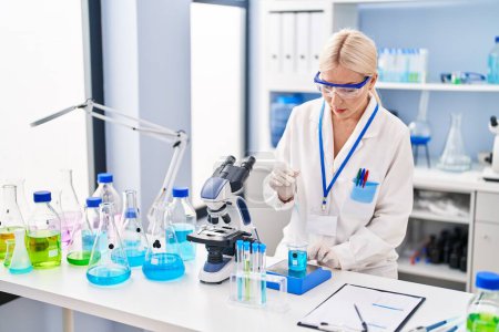Téléchargez les photos : Jeune femme blonde portant un uniforme scientifique mesurant le liquide au laboratoire - en image libre de droit