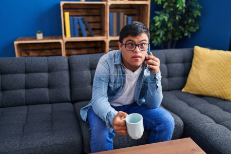 Foto de Síndrome de Down hombre beber café y hablar en el teléfono inteligente en casa - Imagen libre de derechos
