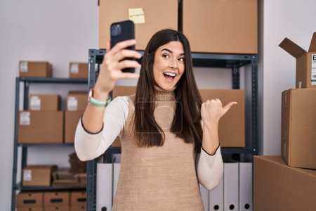 Téléchargez les photos : Jeune femme brune travaillant dans une petite entreprise commerce électronique prenant selfie pointant le pouce vers le côté souriant heureux avec la bouche ouverte - en image libre de droit