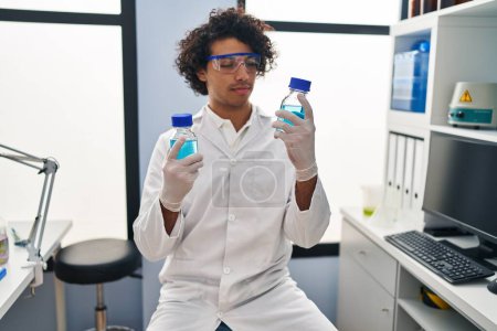 Foto de Joven hispano vistiendo uniforme científico sosteniendo botellas en el laboratorio - Imagen libre de derechos
