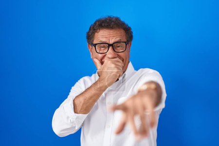 Foto de Middle age hispanic man standing over blue background laughing at you, pointing finger to the camera with hand over mouth, shame expression - Imagen libre de derechos