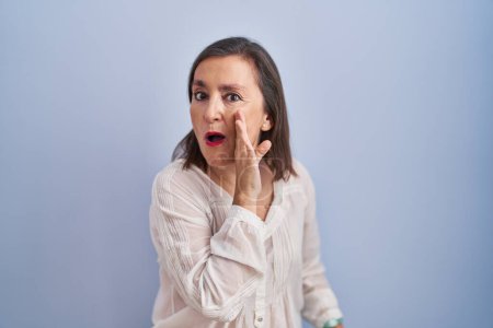 Foto de Mujer hispana de mediana edad de pie sobre fondo azul mano sobre boca contando rumores secretos, susurrando conversación maliciosa - Imagen libre de derechos