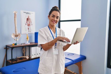 Téléchargez les photos : Young beautiful hispanic woman physiotherapist smiling confident using laptop at rehab clinic - en image libre de droit