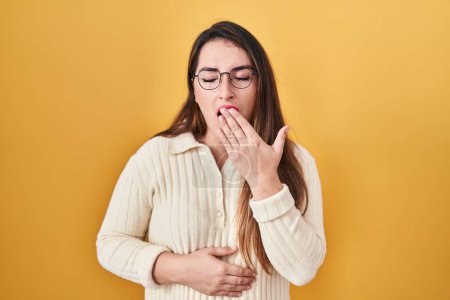 Foto de Mujer hispana joven de pie sobre fondo amarillo aburrida bostezando cansada cubriendo la boca con la mano. inquietud y somnolencia. - Imagen libre de derechos