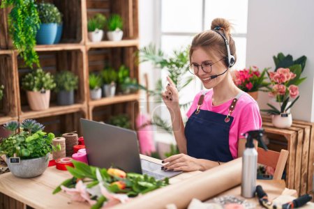 Téléchargez les photos : Blonde femme caucasienne travaillant au magasin de fleuriste faire appel vidéo souriant heureux pointant avec la main et le doigt sur le côté - en image libre de droit