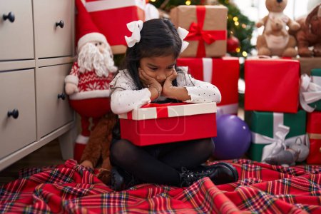 Photo for Adorable hispanic girl holding gift sitting on floor by christmas tree with sad expression at home - Royalty Free Image
