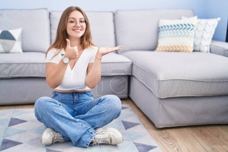 Photo for Young caucasian woman sitting on the floor at the living room showing palm hand and doing ok gesture with thumbs up, smiling happy and cheerful - Royalty Free Image