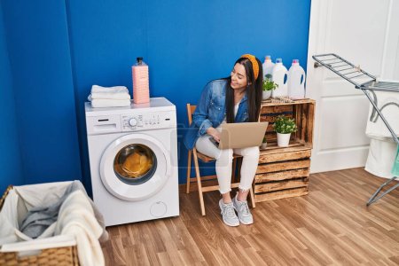 Téléchargez les photos : Young beautiful hispanic woman using laptop waiting for washing machine at laundry room - en image libre de droit