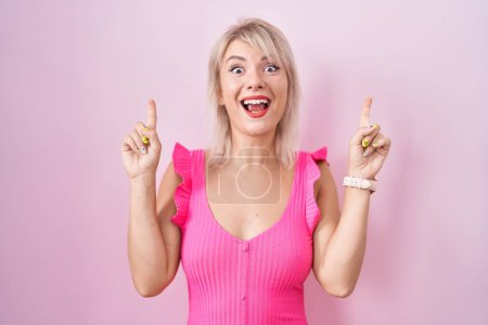Foto de Joven mujer caucásica de pie sobre fondo rosa sonriendo asombrada y sorprendida y señalando con los dedos y los brazos levantados. - Imagen libre de derechos