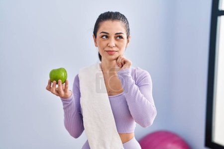 Téléchargez les photos : Jeune femme portant des vêtements de sport et serviette manger pomme verte visage sérieux penser à la question avec la main sur le menton, réfléchi à l'idée déroutante - en image libre de droit