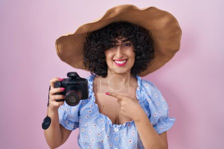 Foto de Young brunette woman with curly hair using reflex camera smiling happy pointing with hand and finger - Imagen libre de derechos