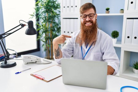 Téléchargez les photos : Homme rousse à longue barbe portant l'uniforme de médecin travaillant à l'aide d'un ordinateur portable souriant heureux pointant avec la main et le doigt - en image libre de droit