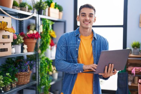Téléchargez les photos : Jeune homme hispanique fleuriste souriant confiant en utilisant un ordinateur portable au magasin de fleuriste - en image libre de droit