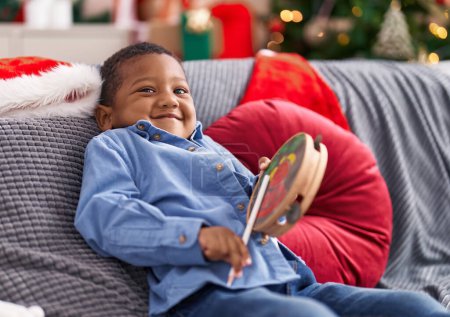 Foto de Niño afroamericano jugando pandereta sentado en el sofá por el árbol de Navidad en casa - Imagen libre de derechos