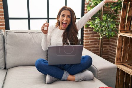 Téléchargez les photos : Jeune femme brune à l'aide d'un ordinateur portable tenant les clés de la nouvelle maison célébrant la victoire avec un sourire heureux et l'expression gagnante avec les mains levées - en image libre de droit