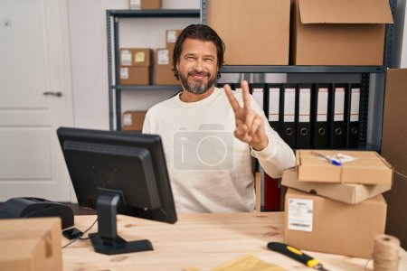 Photo for Handsome middle age man working at small business ecommerce smiling looking to the camera showing fingers doing victory sign. number two. - Royalty Free Image