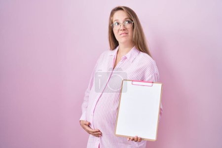 Téléchargez les photos : Jeune femme enceinte tenant presse-papiers souriant regardant vers le côté et regardant loin en pensant. - en image libre de droit