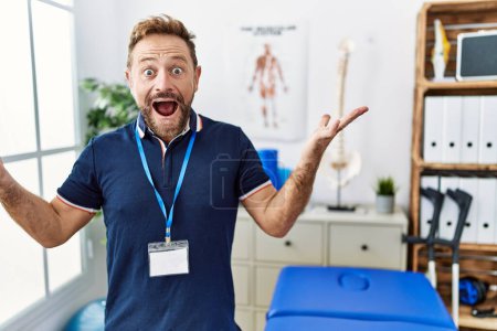 Téléchargez les photos : Middle age physiotherapist man working at pain recovery clinic celebrating crazy and amazed for success with arms raised and open eyes screaming excited. winner concept - en image libre de droit