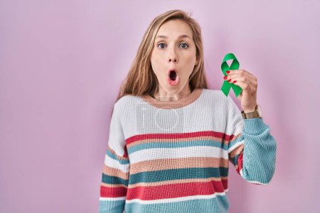 Foto de Joven mujer rubia sosteniendo apoyo cinta verde asustada y sorprendida con la boca abierta para sorpresa, cara de incredulidad - Imagen libre de derechos