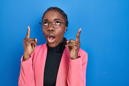Téléchargez les photos : Beautiful black woman standing over blue background amazed and surprised looking up and pointing with fingers and raised arms. - en image libre de droit