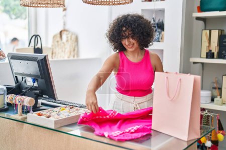 Téléchargez les photos : Young middle eastern woman shop assistant smiling confident folding clothes at clothing store - en image libre de droit