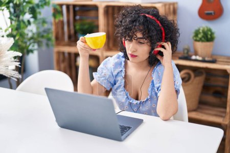 Téléchargez les photos : Young middle east woman using laptop and drinking coffee with serious expression at home - en image libre de droit